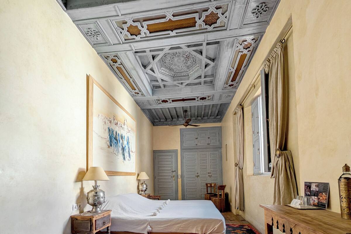 A room with two beds, a ceiling fan, a wooden table, and a window with curtains.