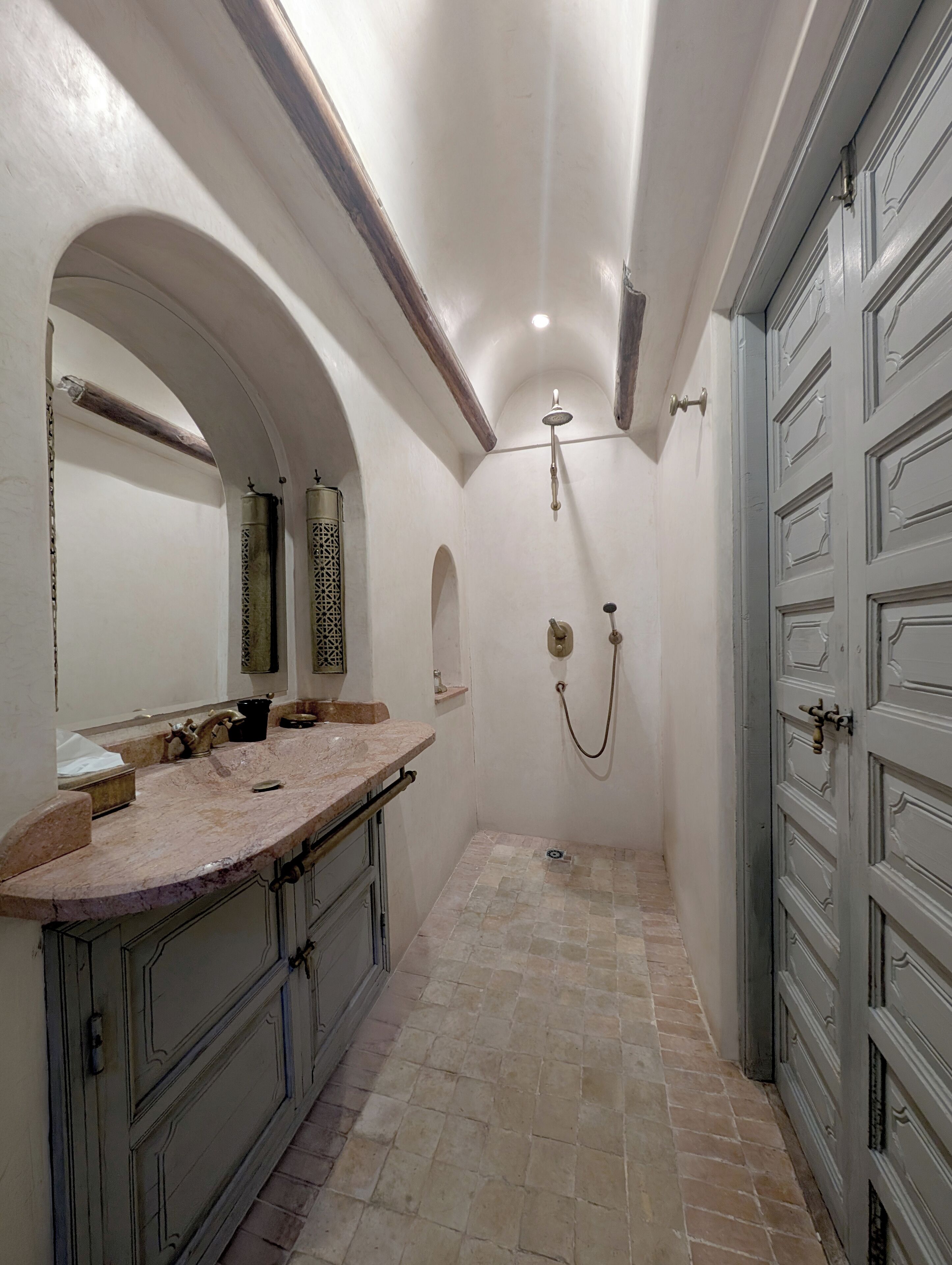 Une petite salle de bain avec un lavabo en marbre, une douche équipée d’un pommeau de douche à main et une grande porte d’entrée en arc de cercle.