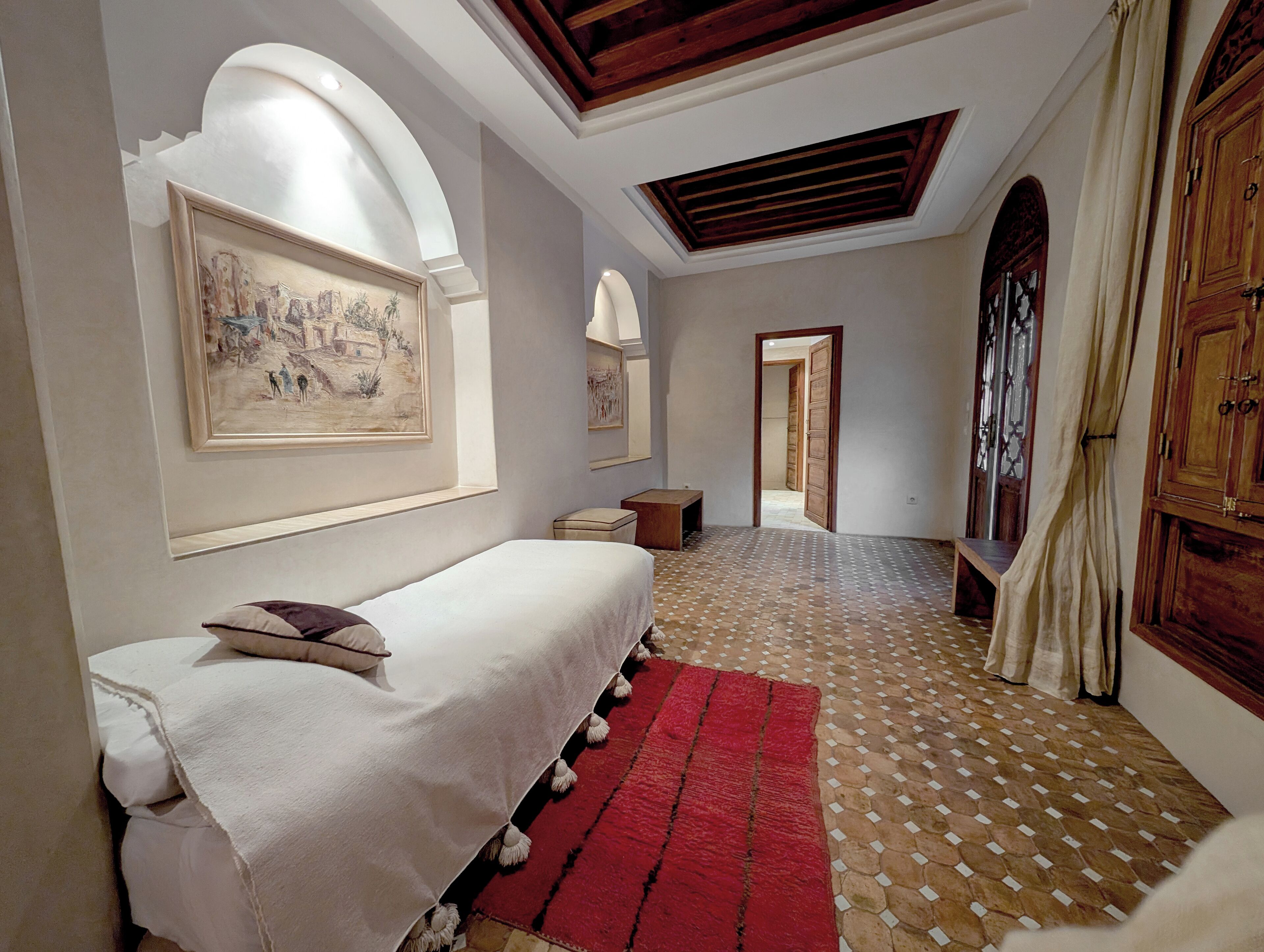 A bedroom with a bed, a red rug, a wooden bench, a painting on the wall, and a wooden door.