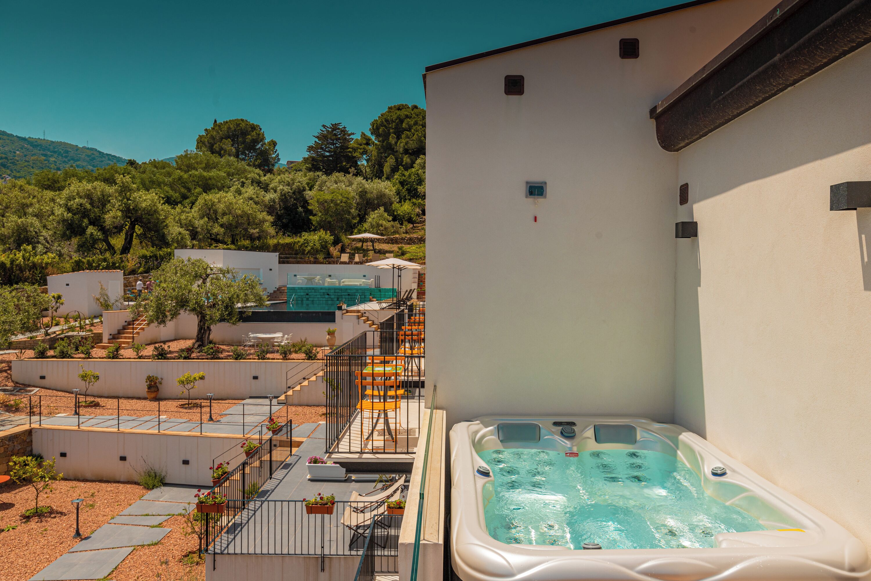 A hot tub adjacent to a white building with a balcony, surrounded by a landscaped garden and a pool area.