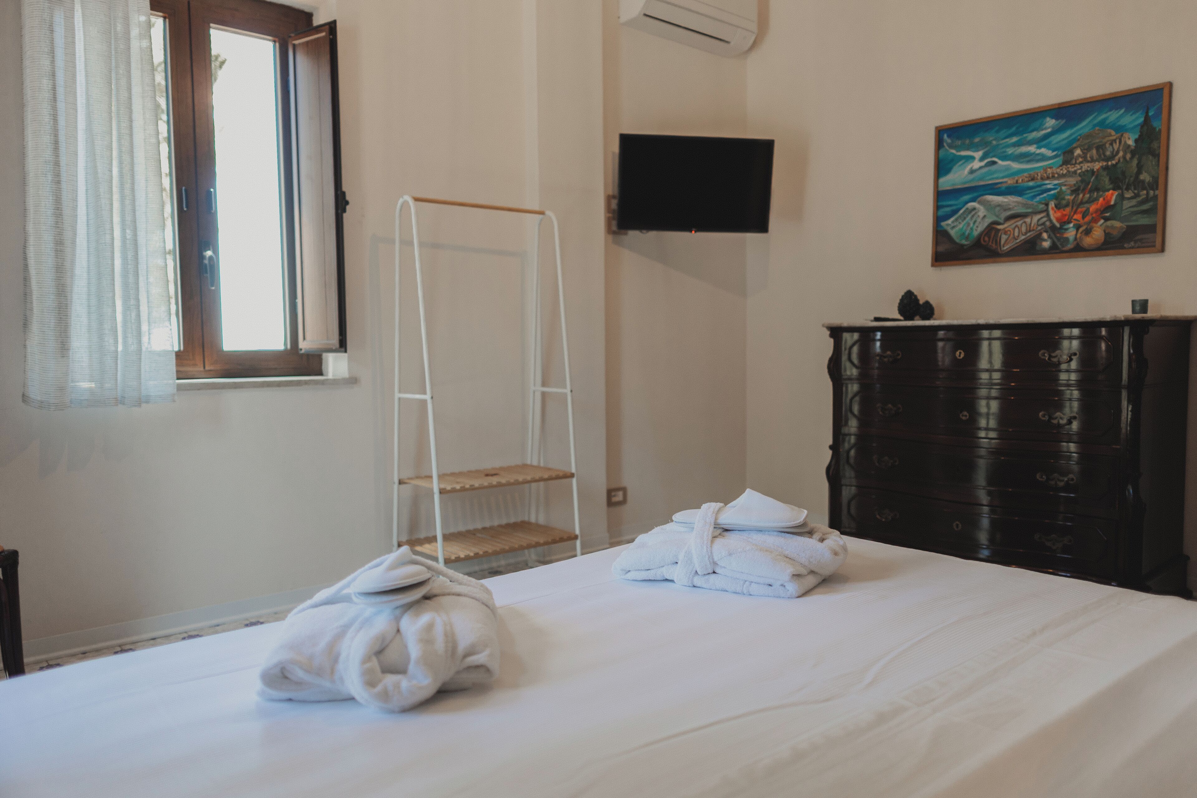 A hotel room with a bed, a white towel on the bed, a wooden dresser, a painting on the wall, and a TV mounted on the wall.