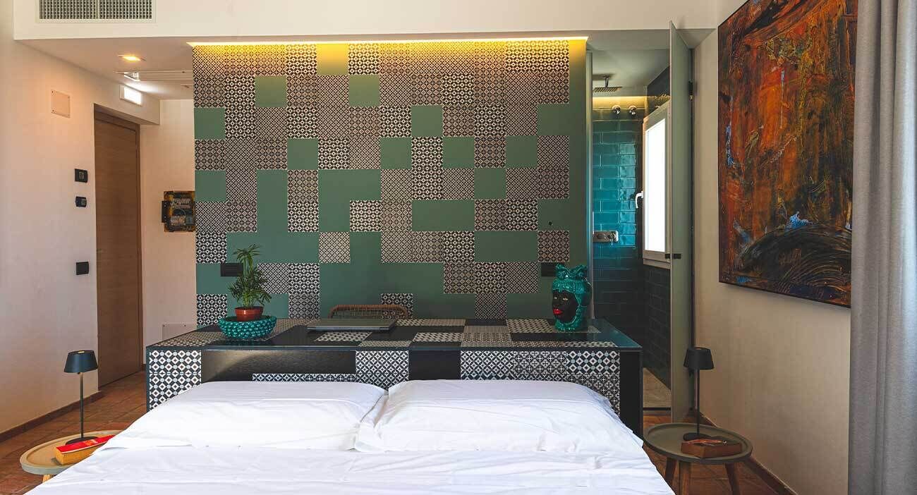 Una habitación de hotel con pared estampada, una cama, una mesita de noche, una planta en maceta y un cuadro.