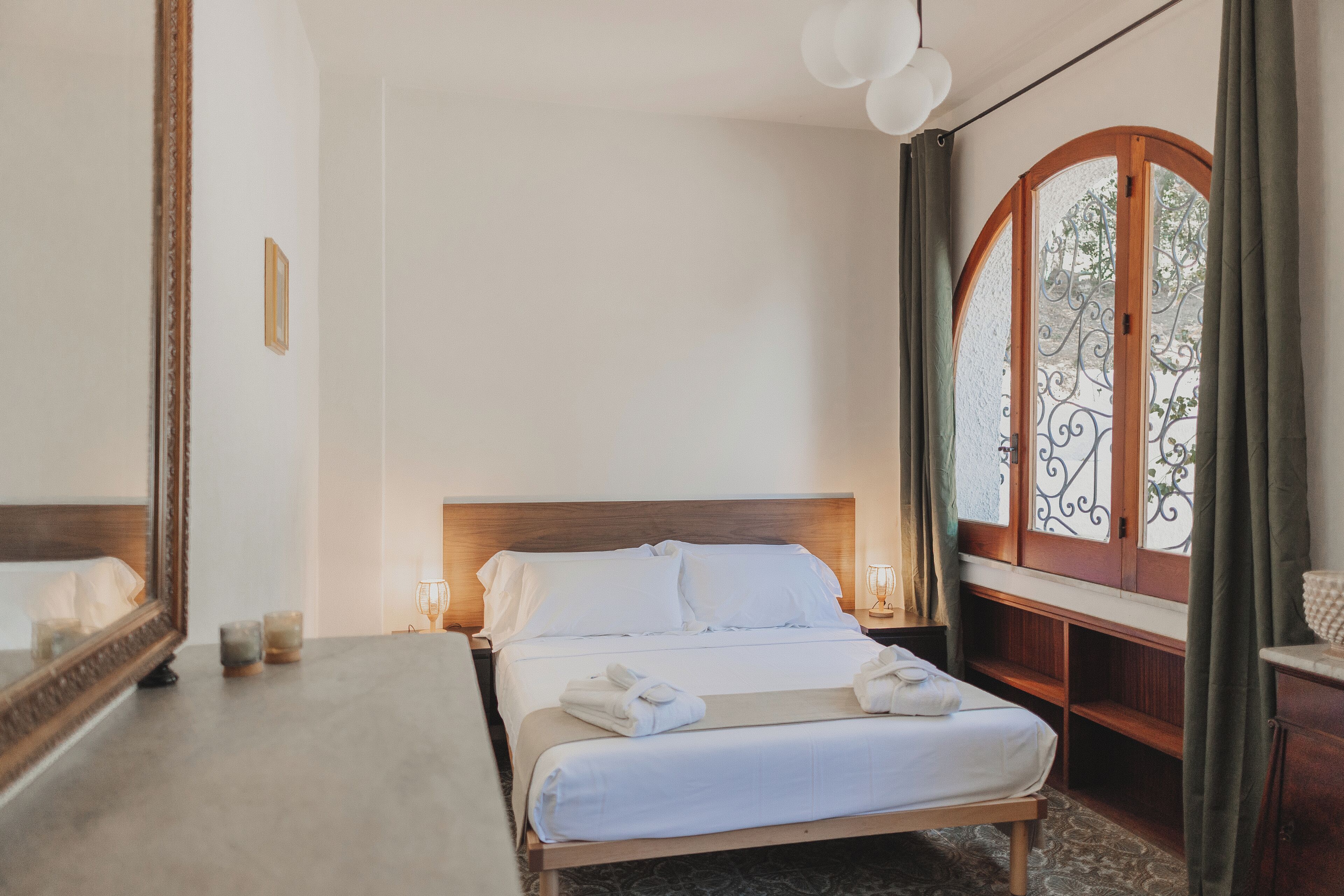 Quarto de hotel com cama, mesas de cabeceira, espelho, janela com cortinas e vista para o verde lá fora.