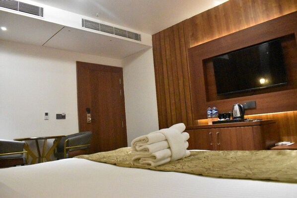 A hotel room with a bed, a TV mounted on the wall, a wooden cabinet, and a desk with a kettle.