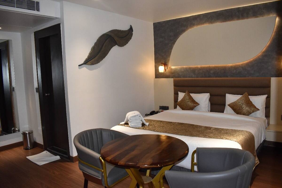 A hotel room with a large bed, a round wooden table, two chairs, a wall-mounted artwork, and a modern light fixture.