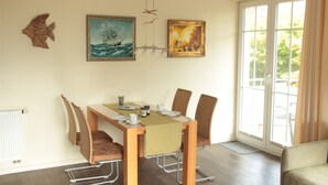 A dining area with a table set for four, surrounded by wicker chairs, with artwork on the walls and a view through a large window. - 4-star apartment near the beach with two balconies (Ostseebad Boltenhagen)
