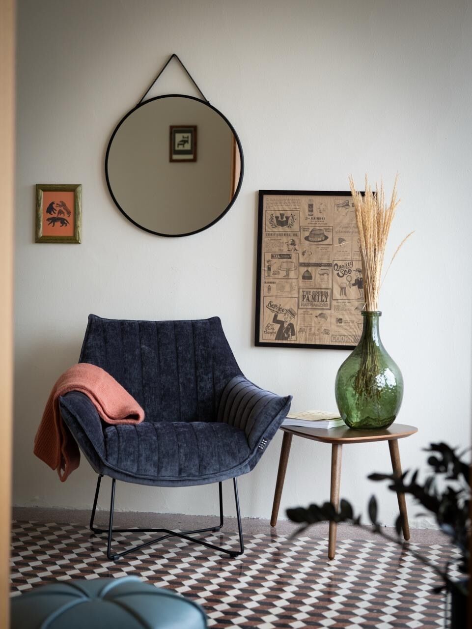 A room with a checkered floor, a blue chair, a small wooden table with a green vase, and a framed poster on the wall.