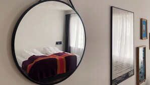 Un espejo circular con marco negro, una cama con sábanas blancas y una manta de colores, y un cuadro en la pared.
