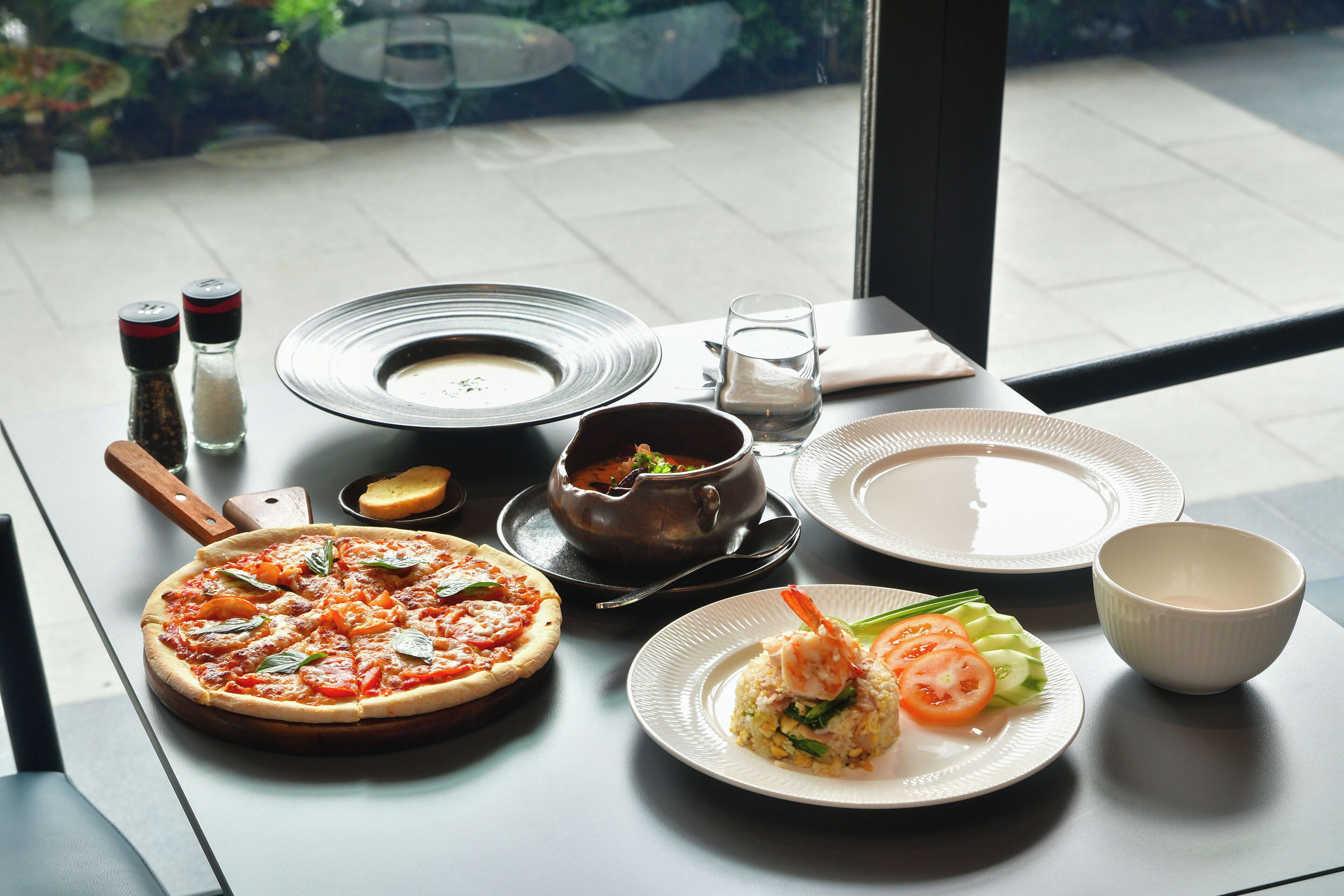 A table with a pizza, soup, rice dish, and plates, set on a table with a window view.
