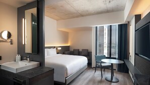 Moderni hotellihuone, jossa on suuri sänky, pyöreä peili, pesuallas ja sen yläpuolella oleva taso, pieni pyöreä pöytä ja televisio.