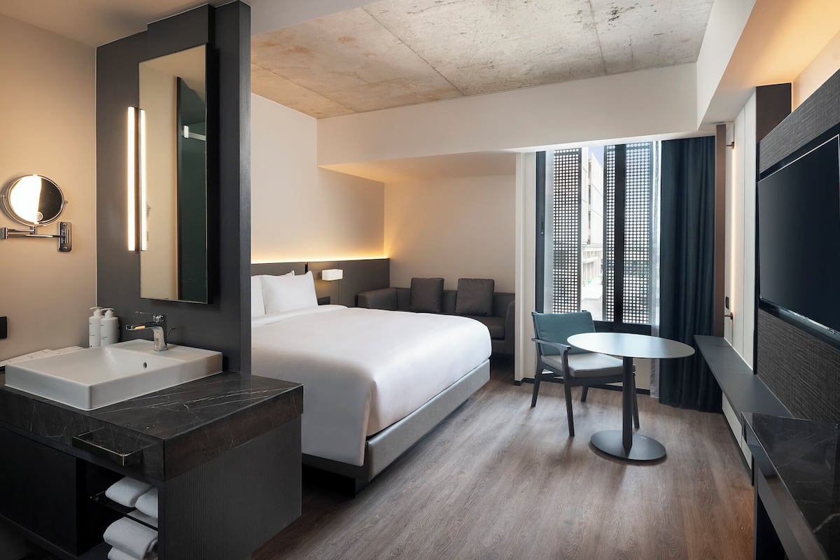 Une chambre d’hôtel moderne avec un grand lit, un miroir rond, une coiffeuse avec un lavabo, une petite table ronde et un téléviseur.