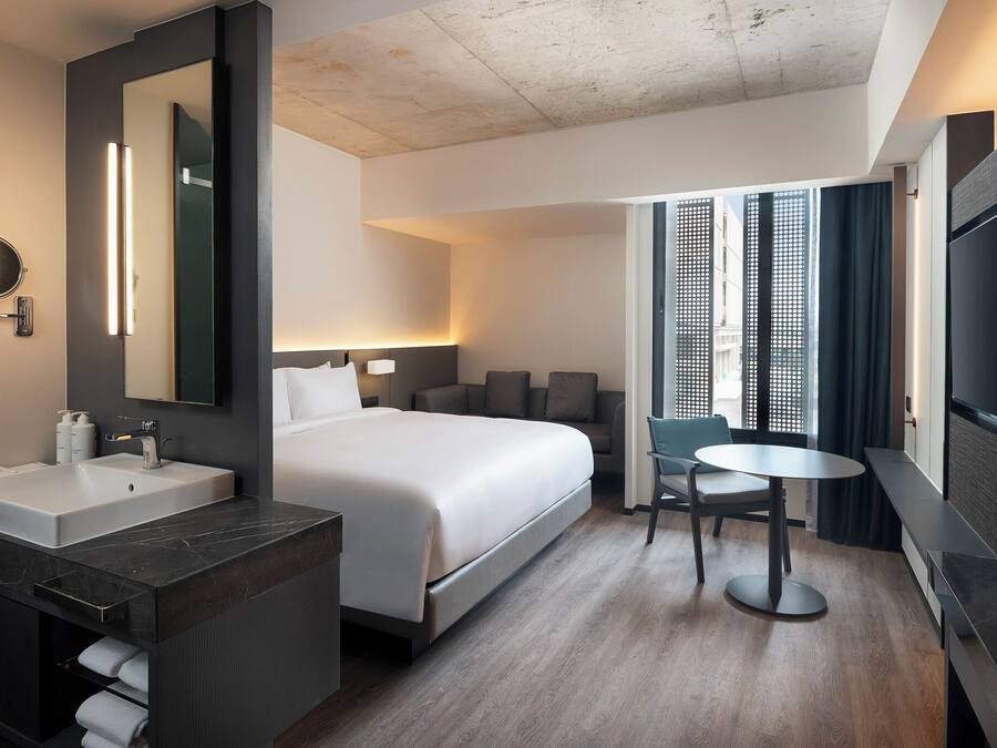 A modern hotel room with a large bed, a round mirror, a vanity with a sink, a small round table, and a TV.