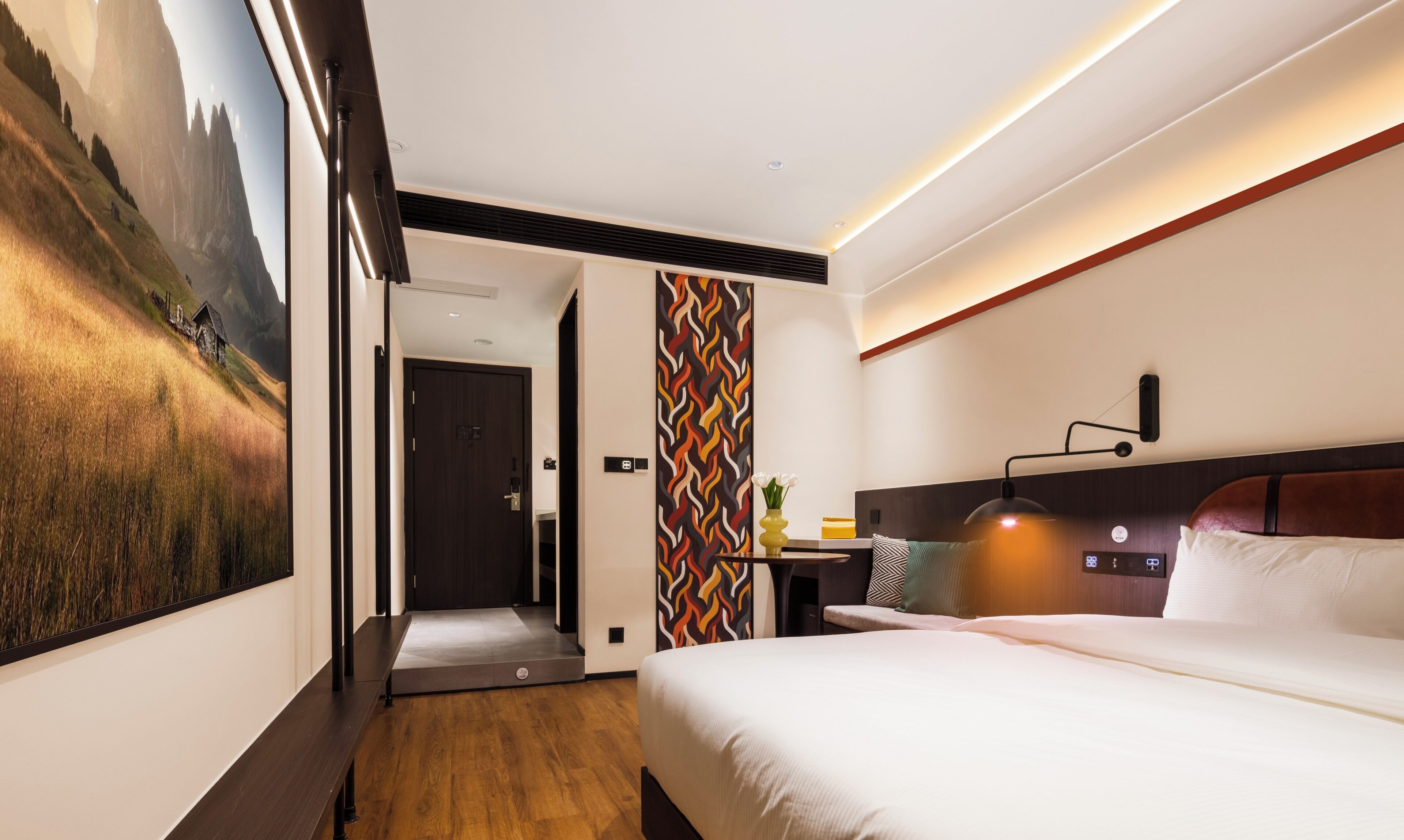 A modern hotel room with a large bed, a desk with a lamp, a view of a mountainous landscape through a large window, and a colorful wall pattern.