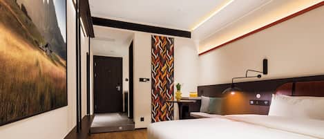 A modern hotel room with a large bed, a desk with a lamp, a view of a mountainous landscape through a large window, and a colorful wall pattern.