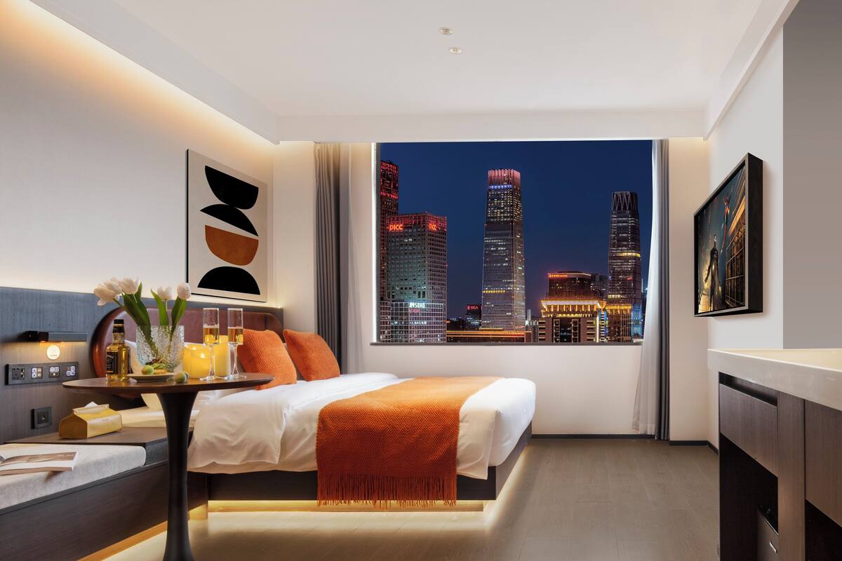 A modern hotel room with a city view, a bed with orange bedding, a bedside table with a lamp, and a large window.