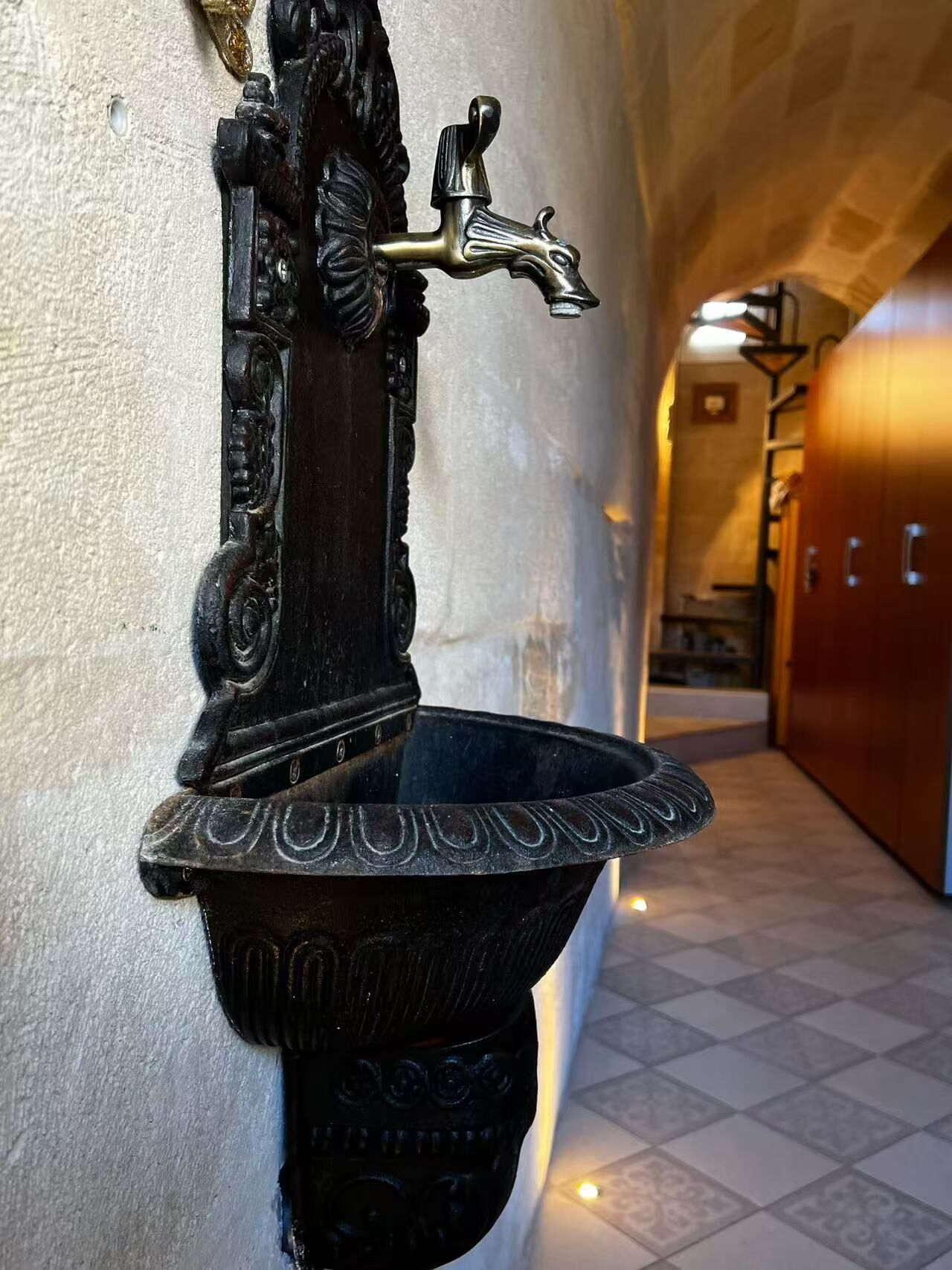 A black, ornate outdoor water fountain with a metal spout and basin, located in a hallway with tiled flooring and wooden doors.