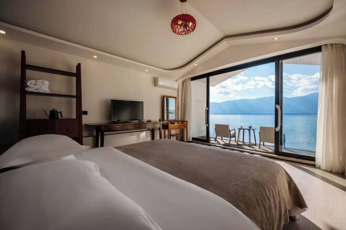 A modern hotel room with a large bed, a desk, a television, and a balcony with a view of the sea.