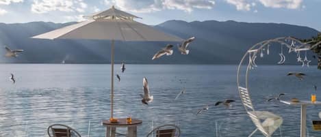 An outdoor seating area with a table and chairs, an umbrella, and a view of a lake with mountains in the background.