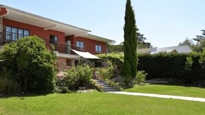 Una casa a due piani con una facciata rossa, un balcone e un giardino curato con prato e un albero alto.