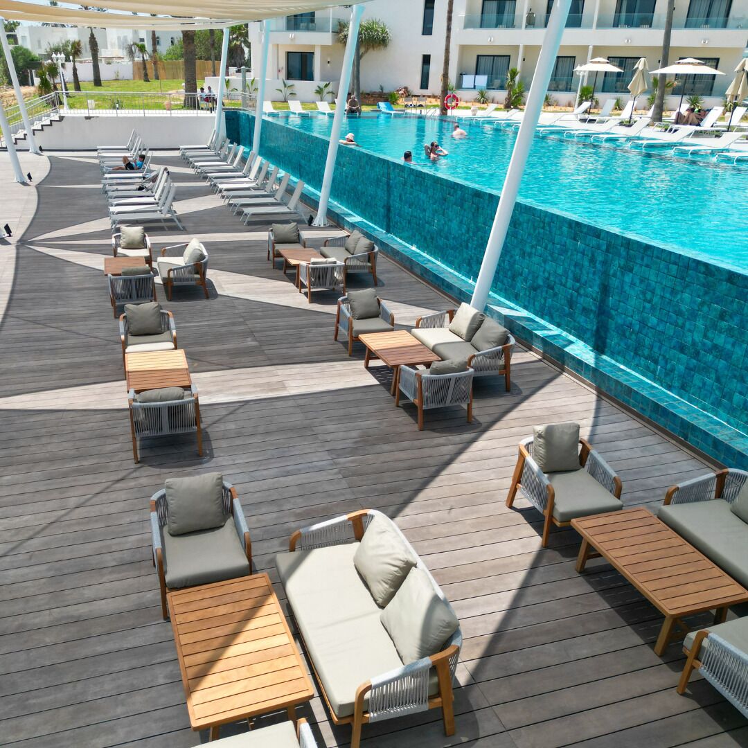 Zona de piscina con sillones y mesas, una piscina azul cristalina y un edificio al fondo.
