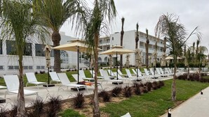 A resort with rows of white lounge chairs and umbrellas, palm trees, and a building in the background. - One Resort Premium  (Hammamet)