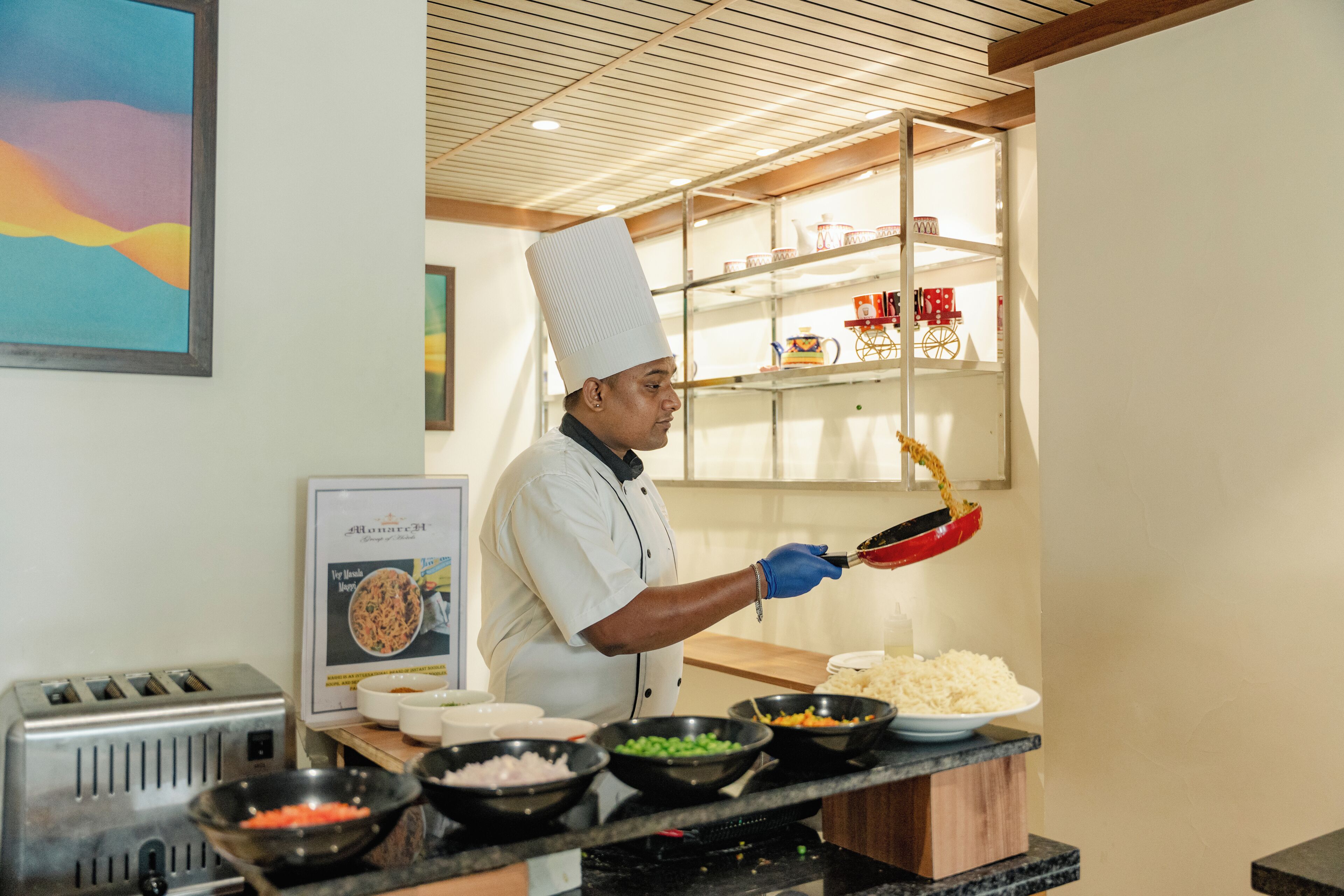 Un chef con uniforme y gorro blanco preparando comida en una cocina con techo y estantes de madera al fondo.