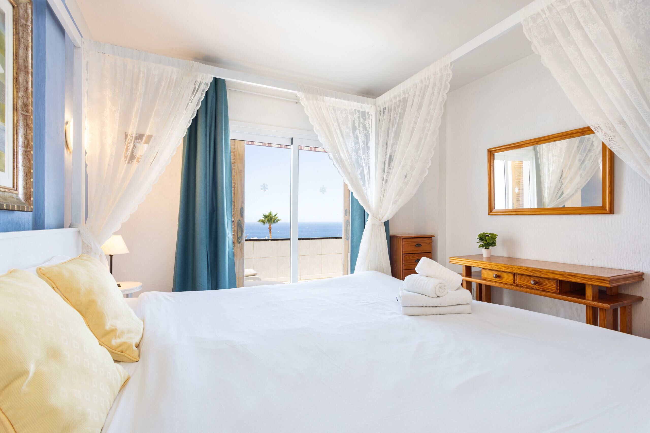 A hotel room with a bed, a wooden dresser, a mirror, and a view of the sea.