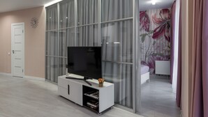 Ett modernt vardagsrum med en stor platt-TV placerad på en vit TV-möbel, en skjutdörr i glas som leder till ett sovrum och en blommig tapet på den intilliggande väggen.