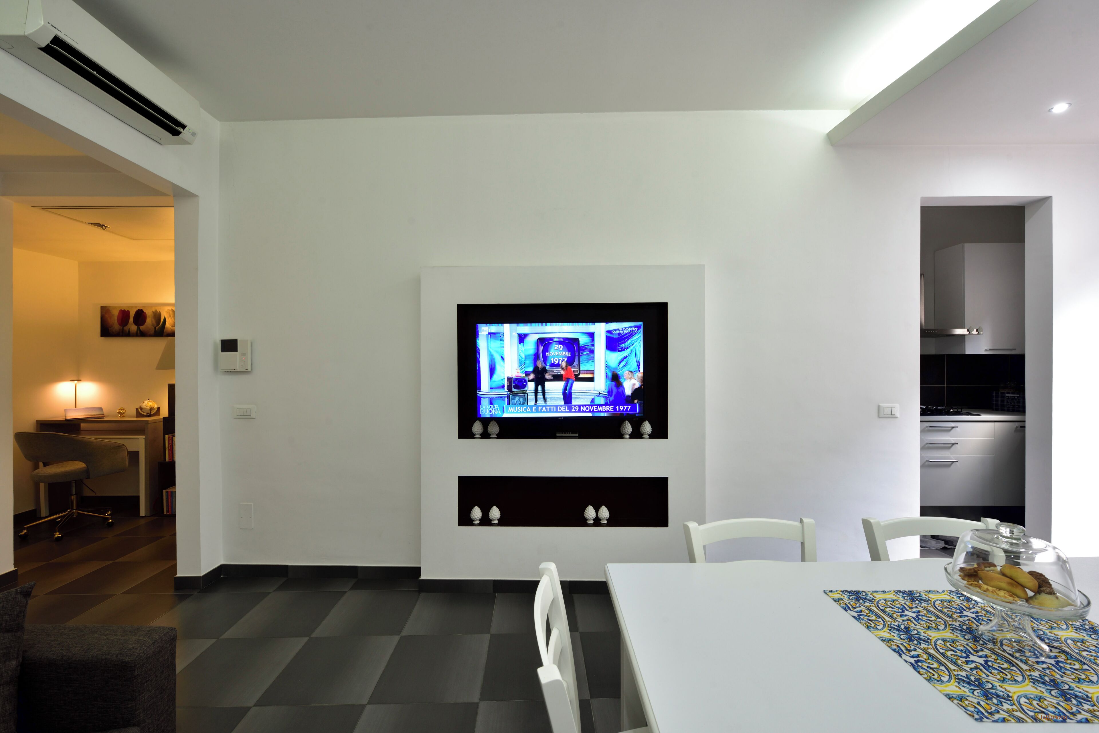 Un espace repas moderne avec un téléviseur fixé au mur, une table blanche et un chemin de table à motifs.