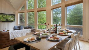 A dining area with a wooden table set for a meal, large windows, and a view of trees.