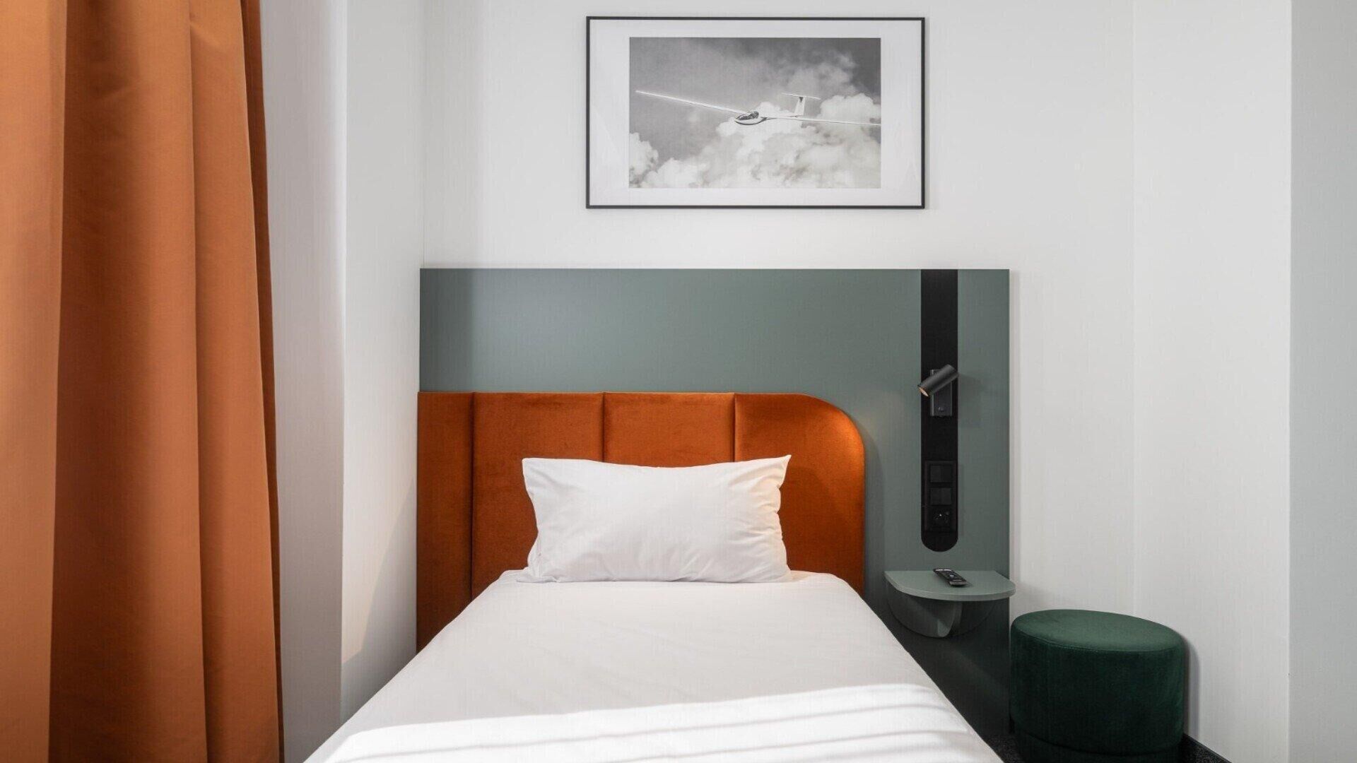 A hotel room with a single bed, a headboard with a grey and orange panel, a bedside table with a remote control, and a green ottoman.