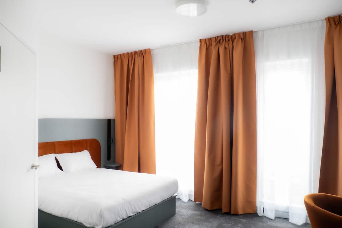Una habitación de hotel moderna con una cama grande, un cabecero gris, una mesita redonda y un sillón marrón.