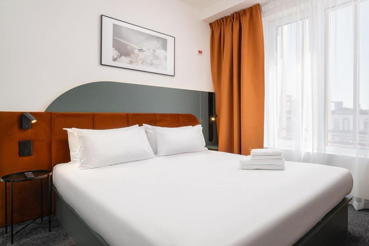 Habitación de hotel con una cama grande, cabecero, mesita de noche, lámpara y ventana con cortinas.