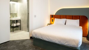 Hotelkamer met een bed, nachtkastje, een wandlamp en een badkamer met wastafel en handdoekrek.