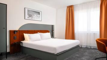 A hotel room with a bed, a nightstand, a chair, a framed picture on the wall, and a window with curtains.