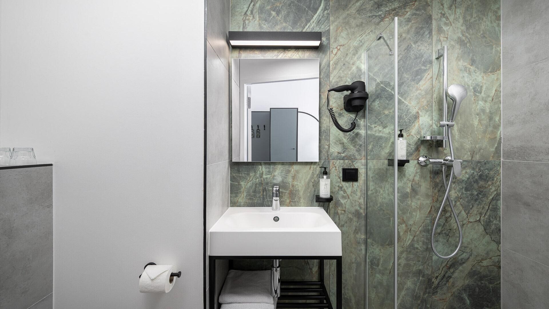 Un baño moderno con un lavabo blanco, un espejo, un perchero para toallas, un sujetapapeles higiénicos y una ducha con cabezal de ducha manual.