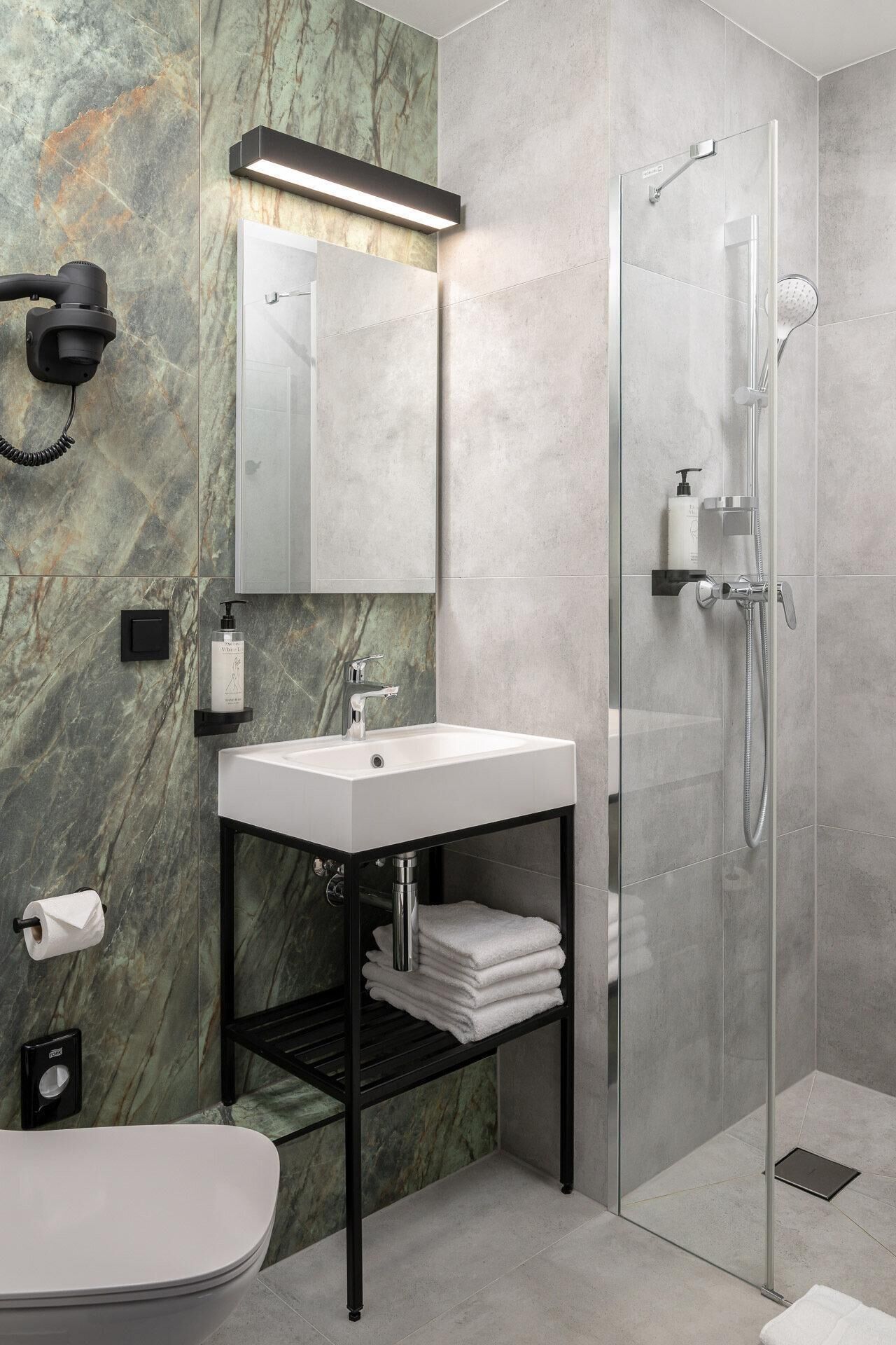 A modern bathroom with a marble wall, a white sink, a glass shower enclosure, and a black shelf with towels.