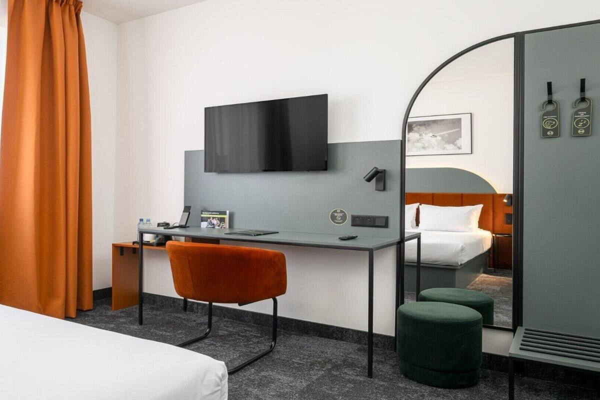 Habitación de hotel con cama, escritorio con silla, televisor, espejo y un otoman verde.