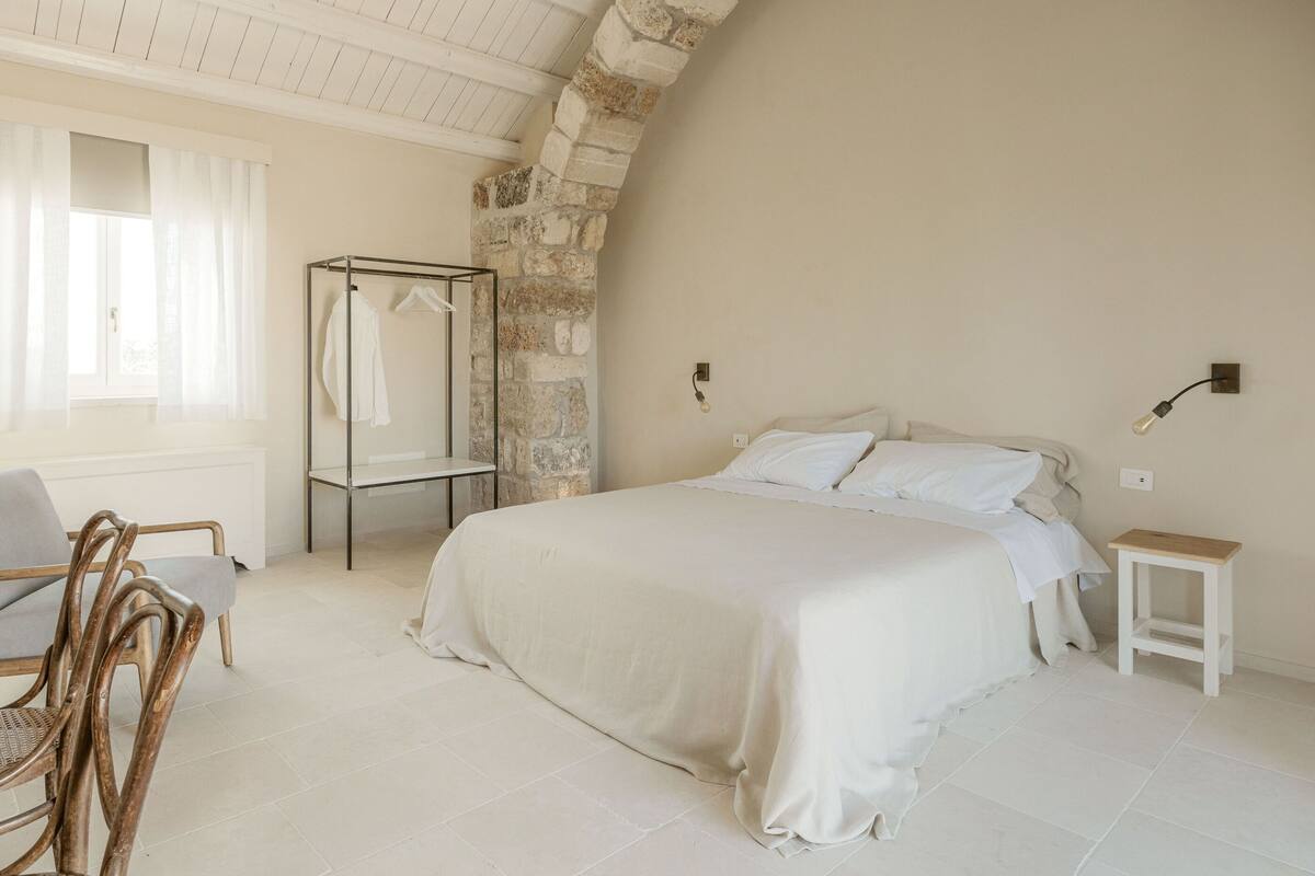 A bedroom with a large bed, a wooden chair, a glass shower enclosure, and a stone wall.