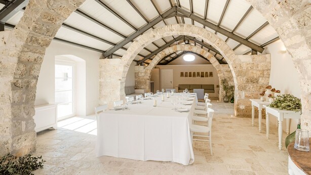 Un comedor amplio con una larga mesa dispuesta para una comida, arcos de piedra y un techo alto con vigas de metal.