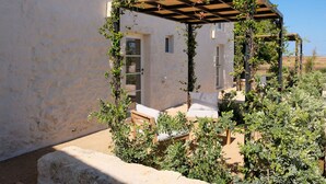 Un espacio de estar al aire libre cubierto, con mobiliario de madera y una pérgola, rodeado de vegetación y un edificio blanco.