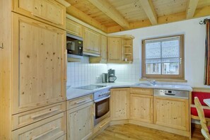 A kitchen with wooden cabinets, a tiled backsplash, a window with blinds, and a red chair. - Scenic Valley Chalet in Austria (Wald im Pinzgau)