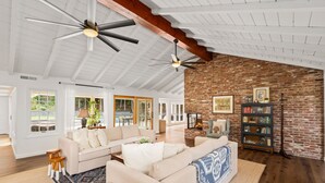 Un salon spacieux avec un plafond haut, des poutres apparentes, un mur décoratif en briques et une cheminée.