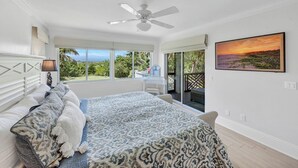 A bedroom with a bed, a ceiling fan, a window with a view, and a painting on the wall.