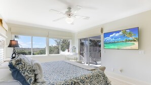 Een slaapkamer met een bed, een plafondventilator, een televisie en een balkon met uitzicht op de oceaan.