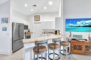 A modern kitchen with a stainless steel refrigerator, a microwave, and a coffee maker. There is a bar area with stools and a large TV screen displaying a tropical beach scene. - Rabbit road oasis just a short stroll to The Gulf! (Sanibel)