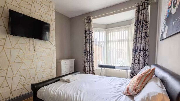 A bedroom with a bed, a flat-screen TV mounted on the wall, a window with curtains, and a patterned wall. - Cornwall House Home from Home Stay (Hartlepool)