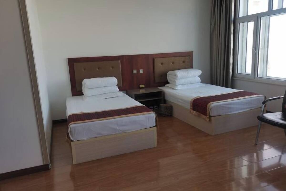 Une chambre d’hôtel avec deux lits simples, une tête de lit en bois, une fenêtre avec des rideaux et une chaise.