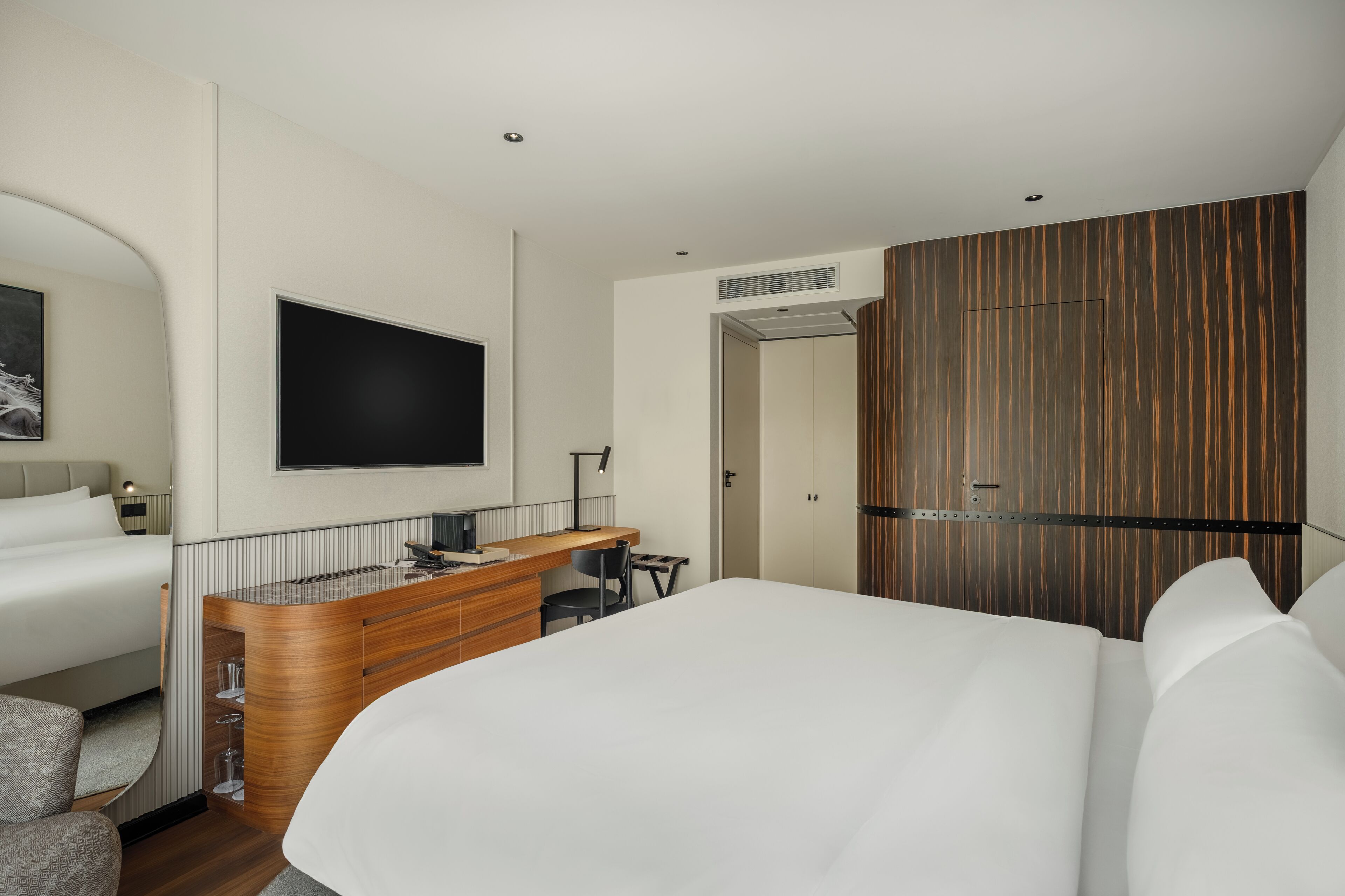 Quarto de hotel moderno com uma cama grande, uma escrivaninha com cadeira, televisão de tela plana fixada na parede e um guarda-roupa com design de painel de madeira.