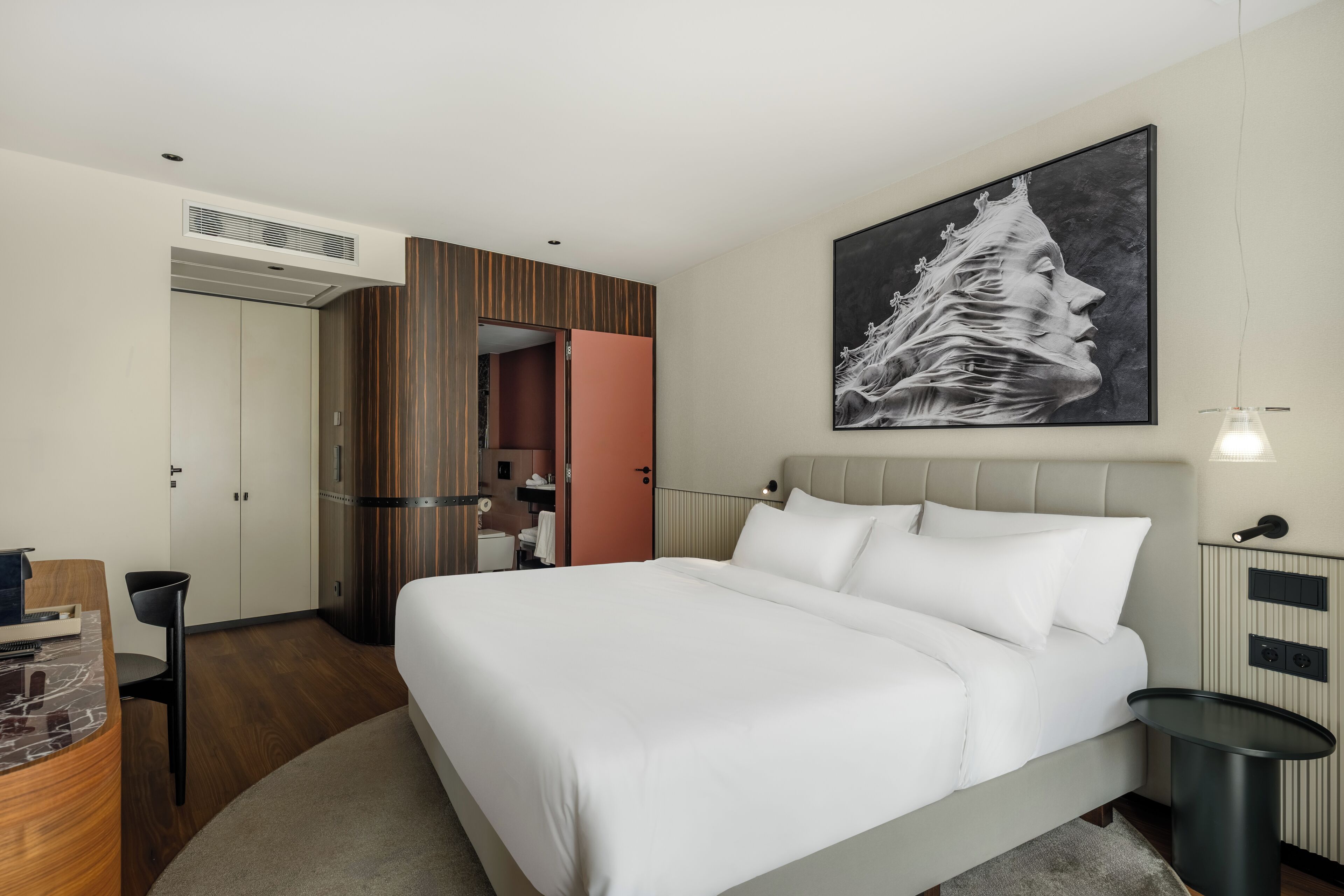 Habitació d'hotel moderna amb un llit gran, una taula de treball i una cadira. Hi ha un quadre a la paret i una finestra amb cortines.
